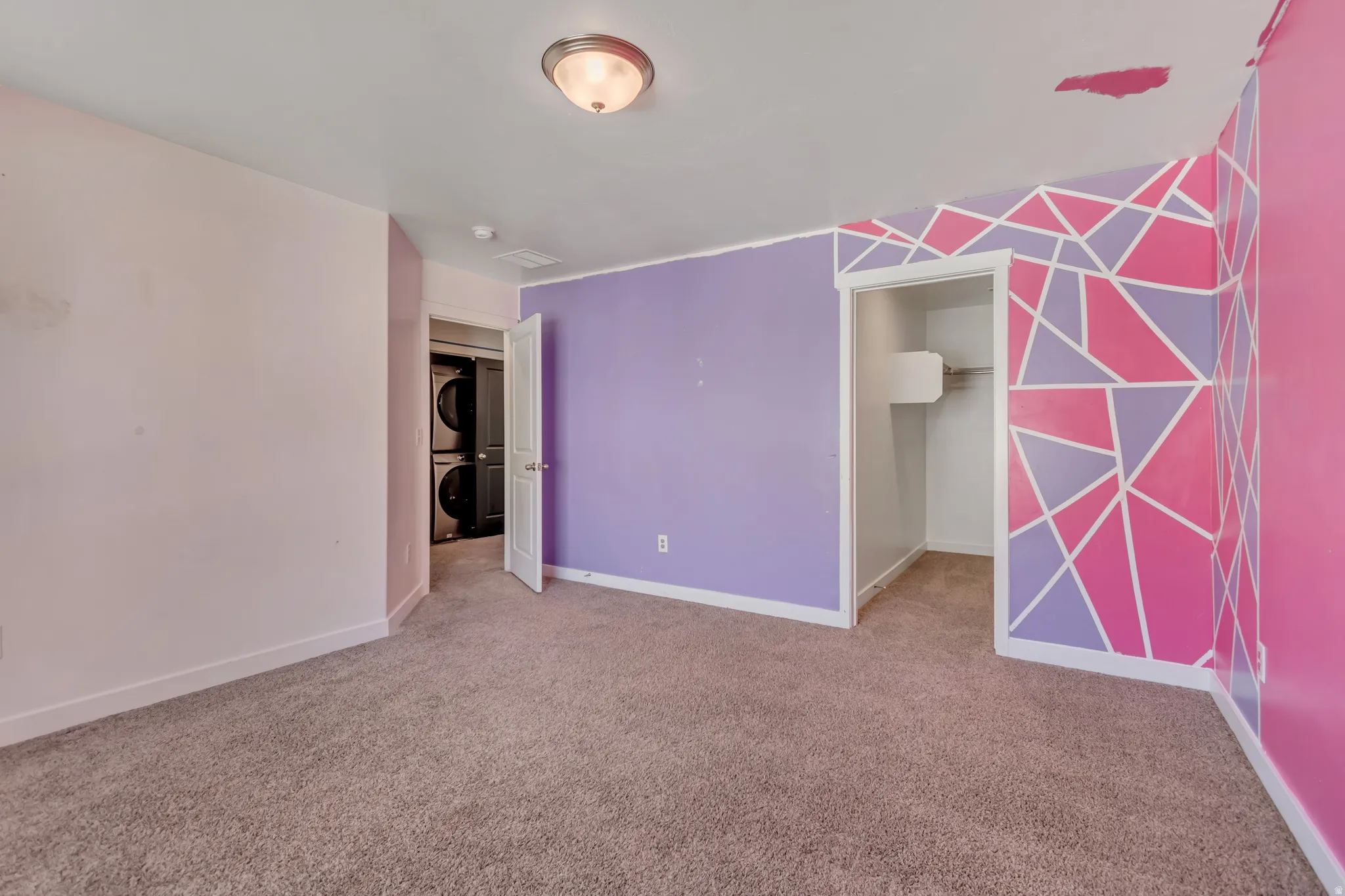 Unfurnished bedroom featuring an accent wall, a walk in closet, stacked washer / drying machine, and light carpet