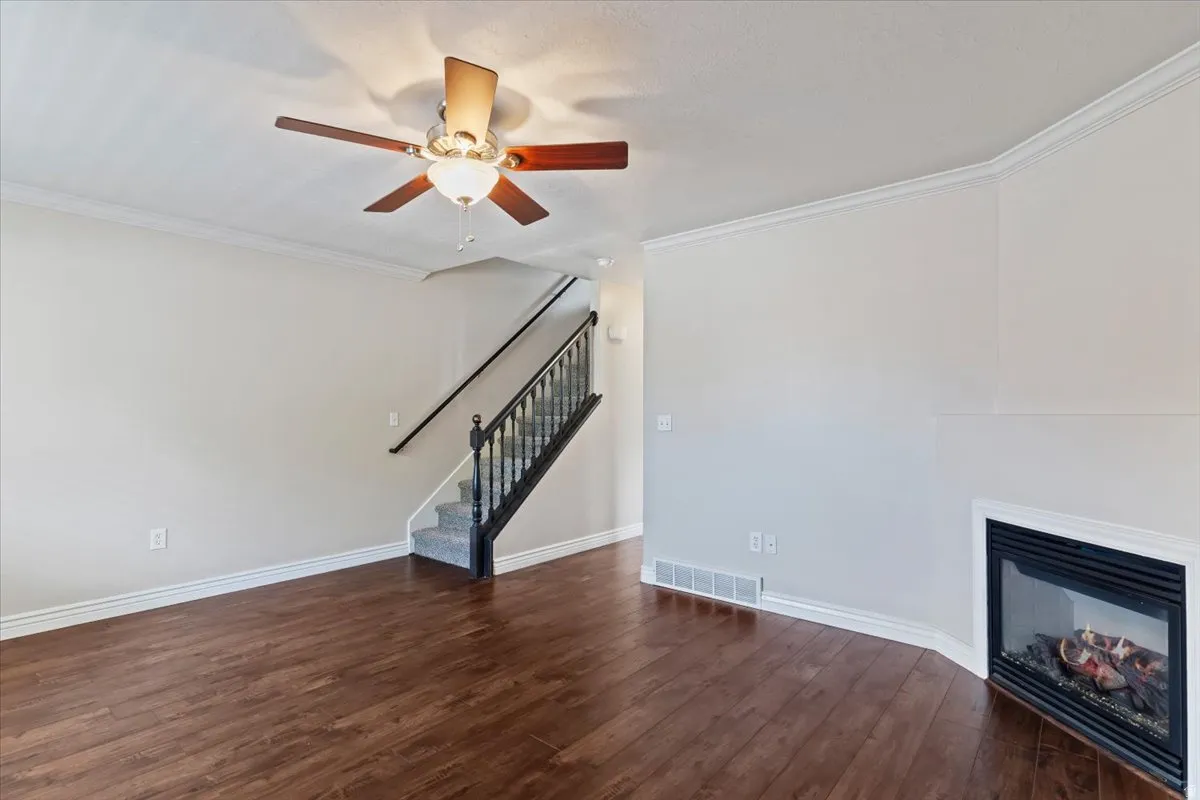 Unfurnished living room featuring a glass covered fireplace, ornamental molding, dark wood-type flooring, and a ceiling fan