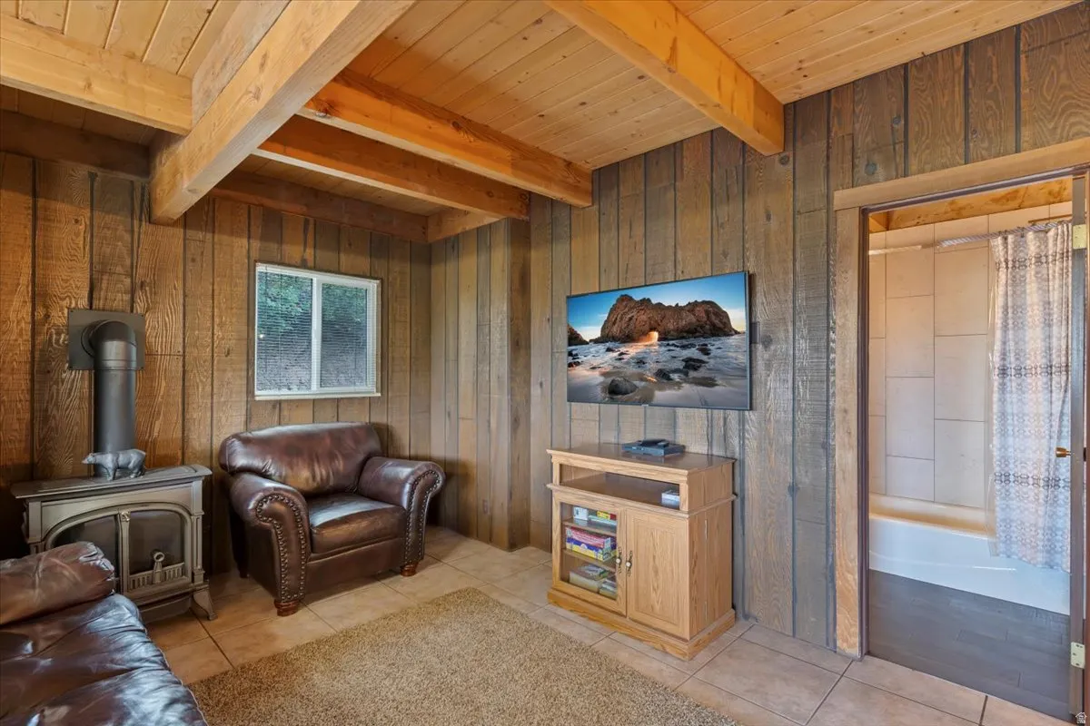 Living room featuring light tile patterned flooring, wood walls, a wood stove, and a wooden ceiling with exposed beams