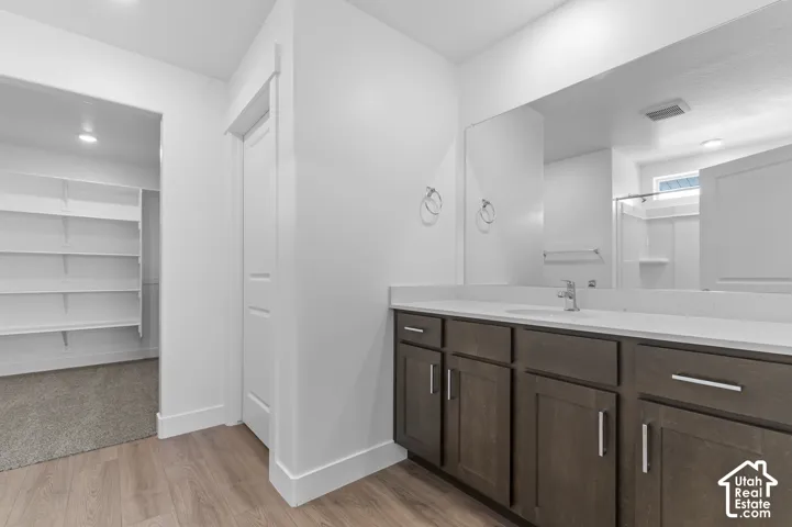 Wood-finish flooring, a vanity with a white countertop and dark wood-finish cabinetry, a large wall-mounted mirror, and an adjacent walk-in closet with extensive shelving