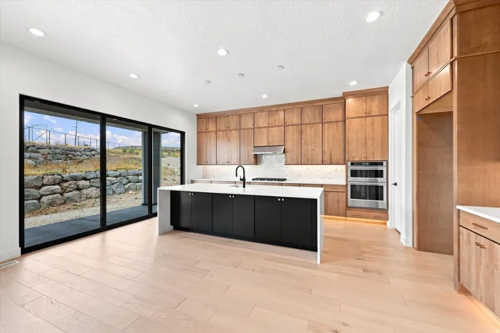 Modern kitchen featuring light wood-finish flooring, a large island with a white countertop and black cabinetry, wood cabinetry, stainless steel double ovens, and expansive sliding glass doors