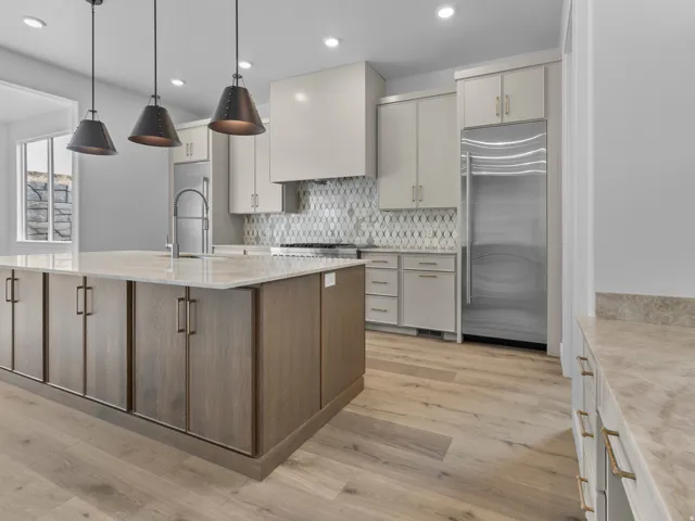 Modern kitchen featuring cream-colored cabinetry with brushed brass hardware, a two-tier island with wood-finish base and white quartz countertop, stainless steel appliances including a refrigerator and dishwasher, geometric tile backsplash, pendant light