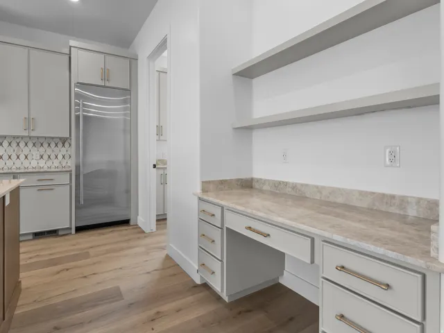 Integrated kitchen desk area featuring a stone-finish countertop, light gray cabinetry with bronze-finish hardware, and open shelving