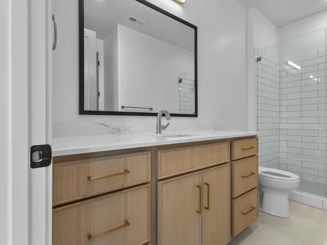 Modern primary bathroom featuring a wood-finish vanity with dual storage drawers and brass hardware, white quartz countertop, rectangular mirror with black frame, polished chrome faucet, and subway tile shower enclosure with glass panel and integrated she