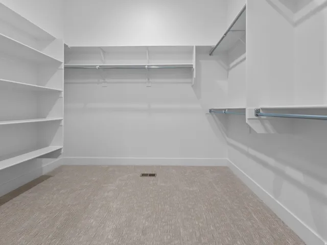 Spacious closet featuring extensive built-in shelving, multiple hanging rods, clean white walls, a visible floor vent, and light-toned patterned carpeting