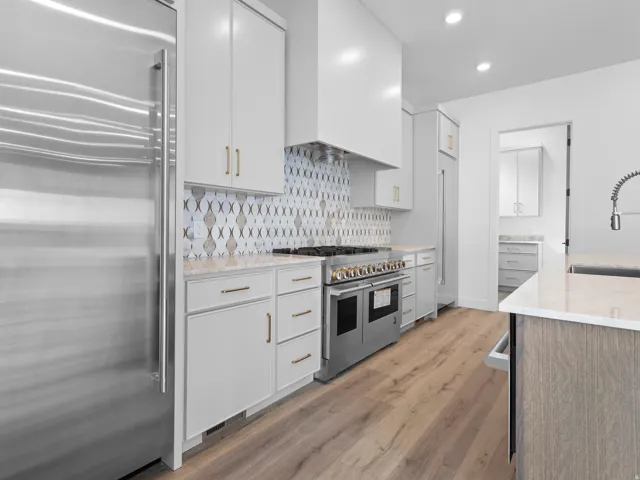 Gourmet kitchen featuring stainless steel appliances, custom range hood, patterned tile backsplash, light wood-finish flooring, and white cabinetry with brass hardware