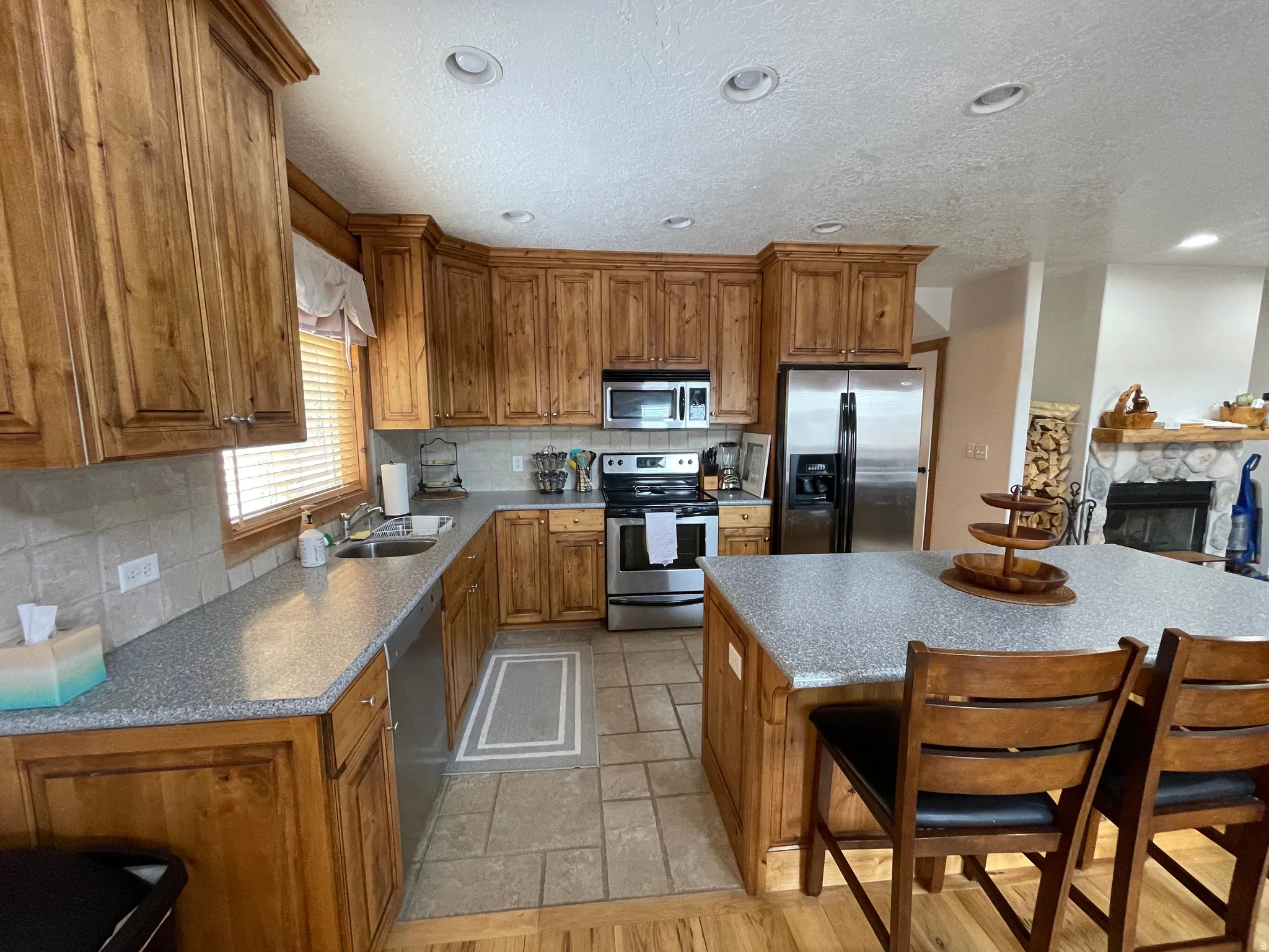 Kitchen featuring backsplash, stainless steel appliances, wood finish cabinets, a stone fireplace, and a textured ceiling