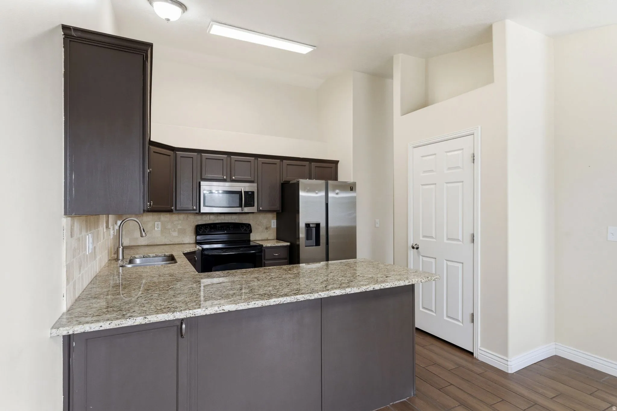 Kitchen with dark wood finish cabinets, stainless steel appliances, a peninsula, light stone counters, and backsplash