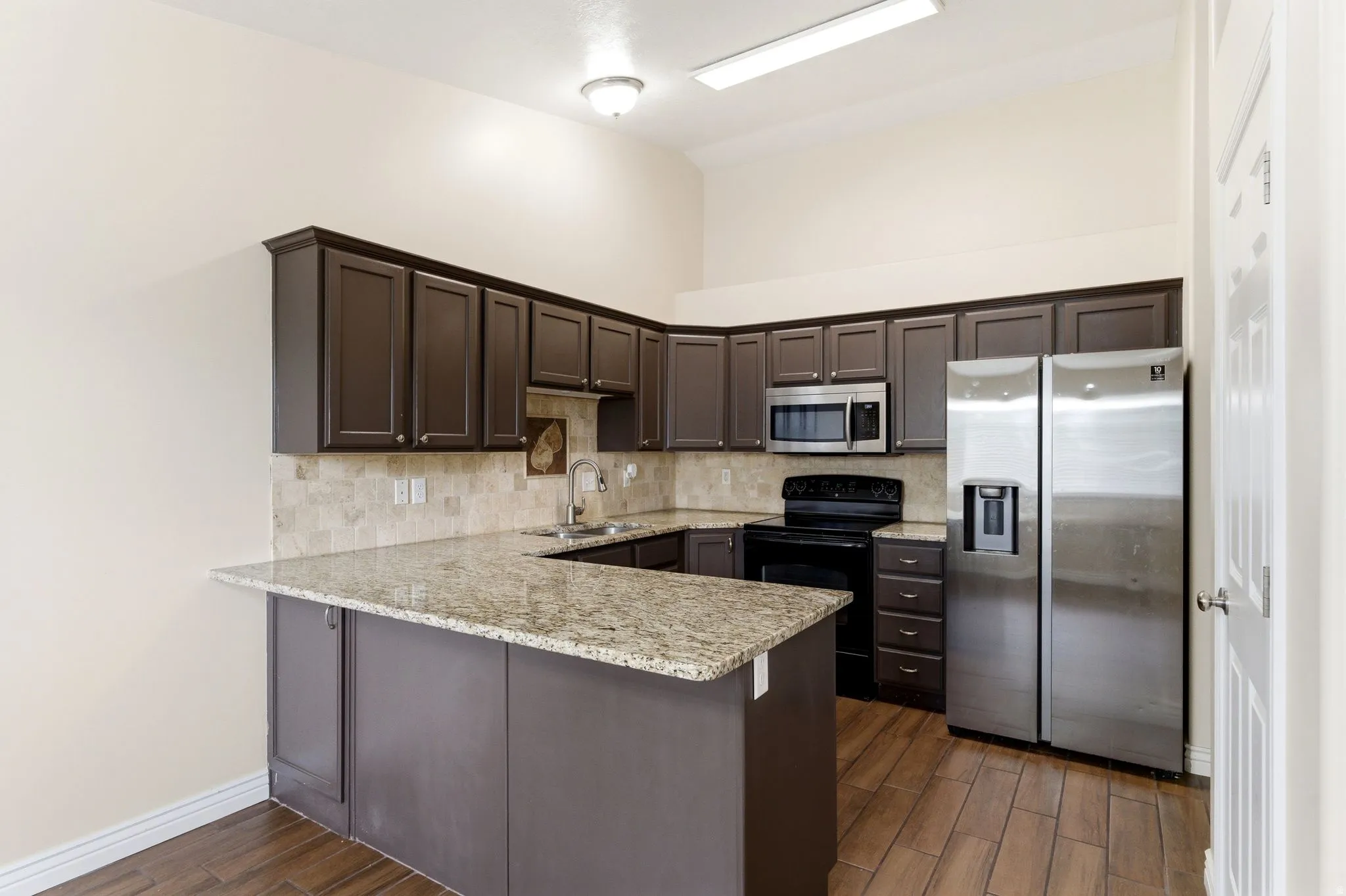 Kitchen featuring dark wood finish cabinets, stainless steel appliances, a peninsula, light stone counters, and dark wood-style flooring