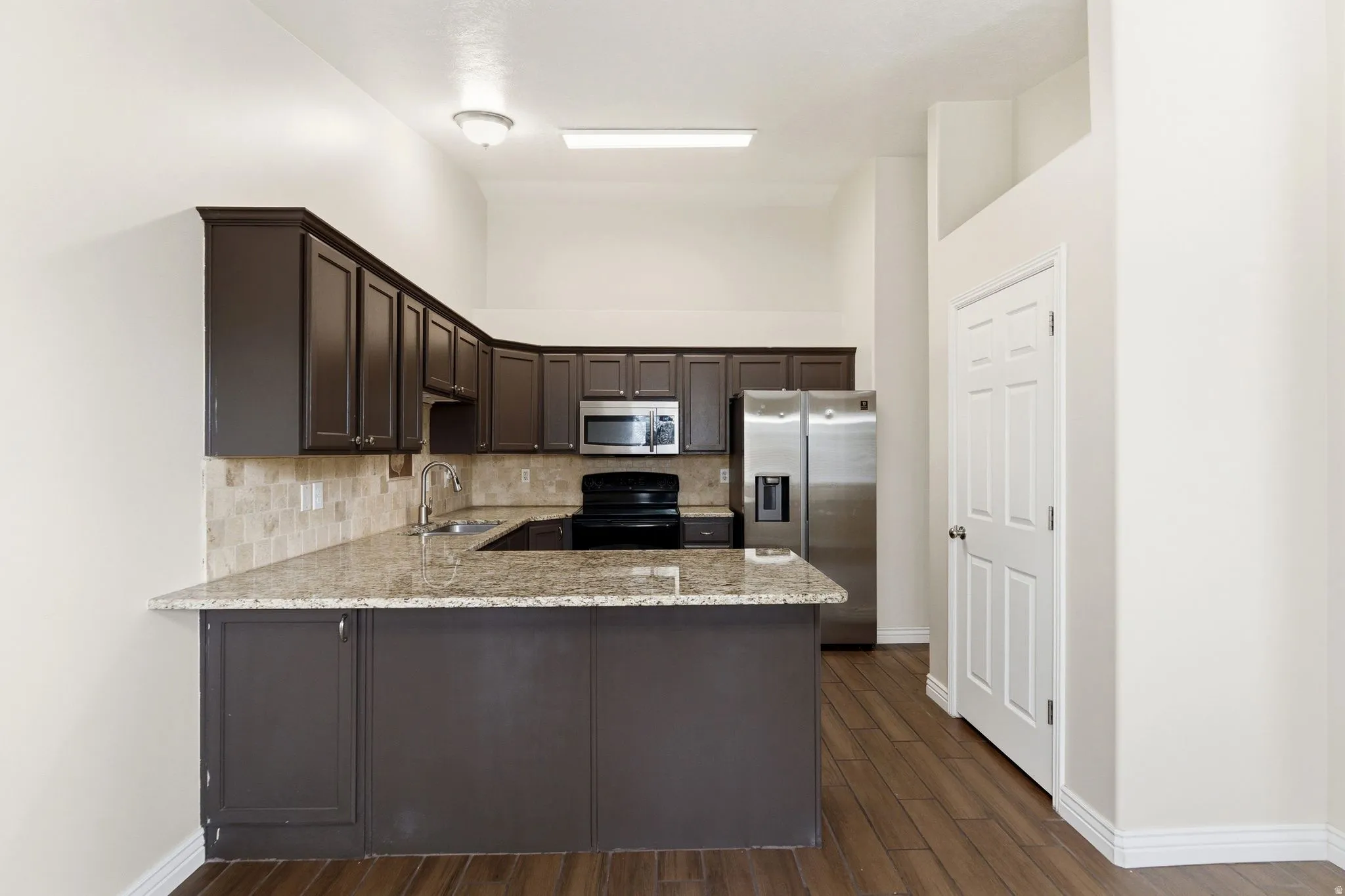 Kitchen with dark wood finish cabinetry, stainless steel appliances, light stone counters, a peninsula, and backsplash