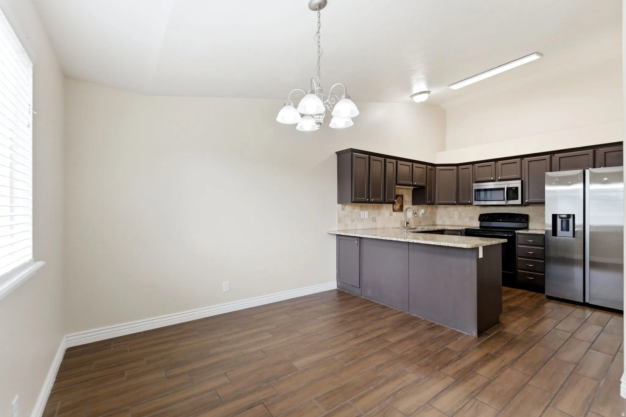Kitchen with stainless steel appliances, a peninsula, suspended lighting, dark wood finish cabinets, and tasteful backsplash
