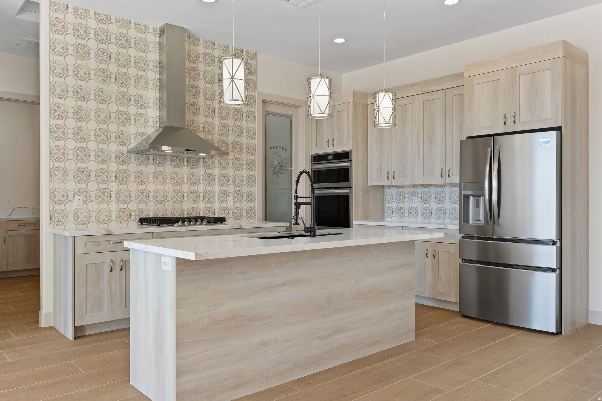 Kitchen with light wood finish cabinetry, stainless steel appliances, backsplash, a center island with sink, and decorative light fixtures