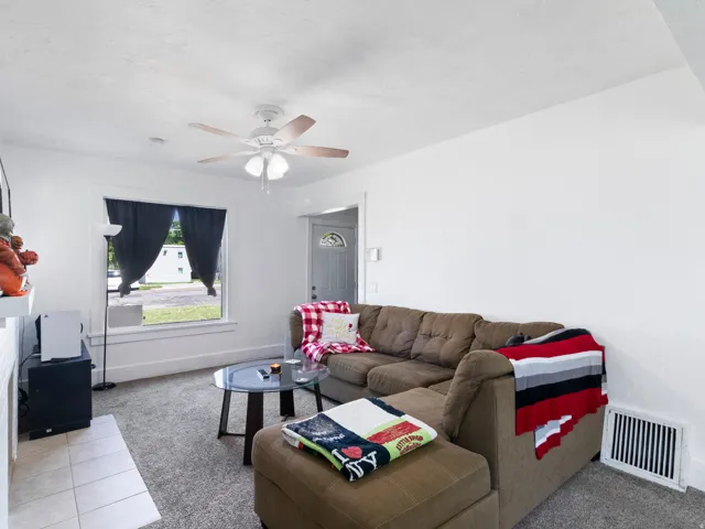 Living area featuring light gray carpeting, white walls, and a ceiling fan with wood-finish blades