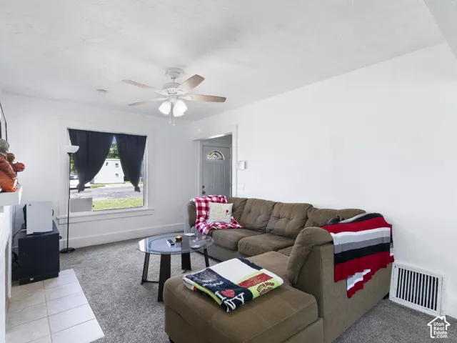 Living area featuring light gray carpeting, white walls, and a ceiling fan with wood-finish blades