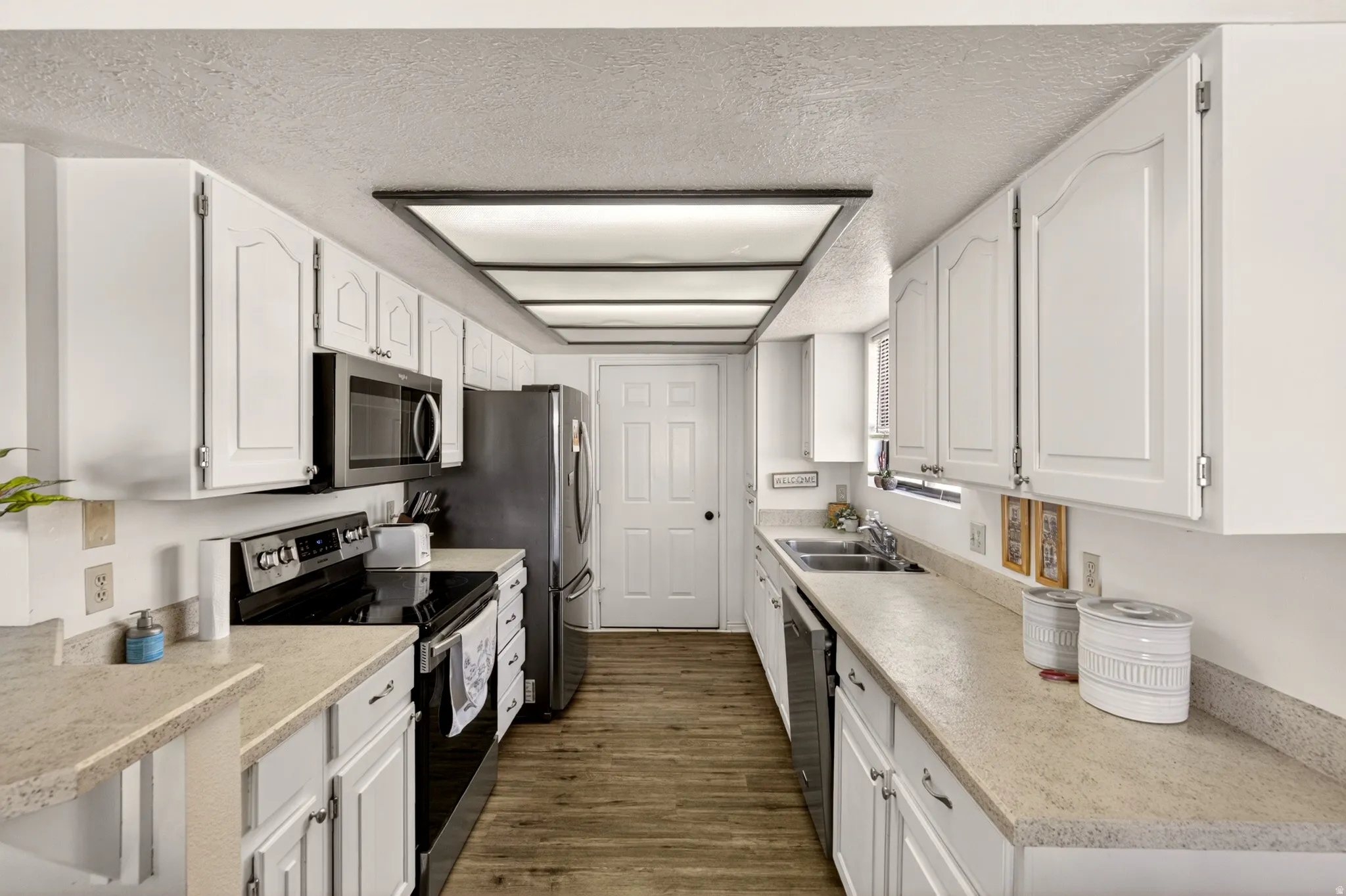 Kitchen with stainless steel appliances, a textured ceiling, white cabinetry, light countertops, and dark wood-style flooring