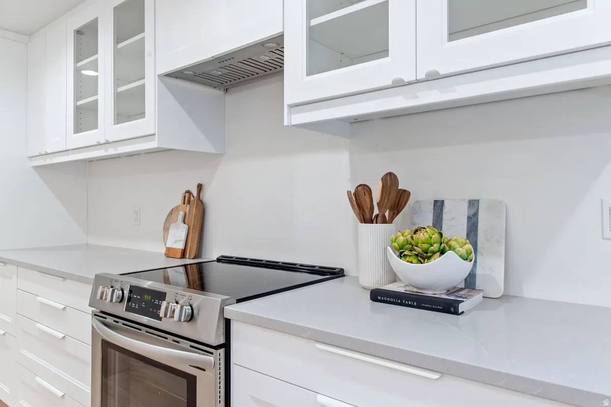 Kitchen featuring glass insert cabinets, electric range, white cabinetry, exhaust hood, and light stone counters