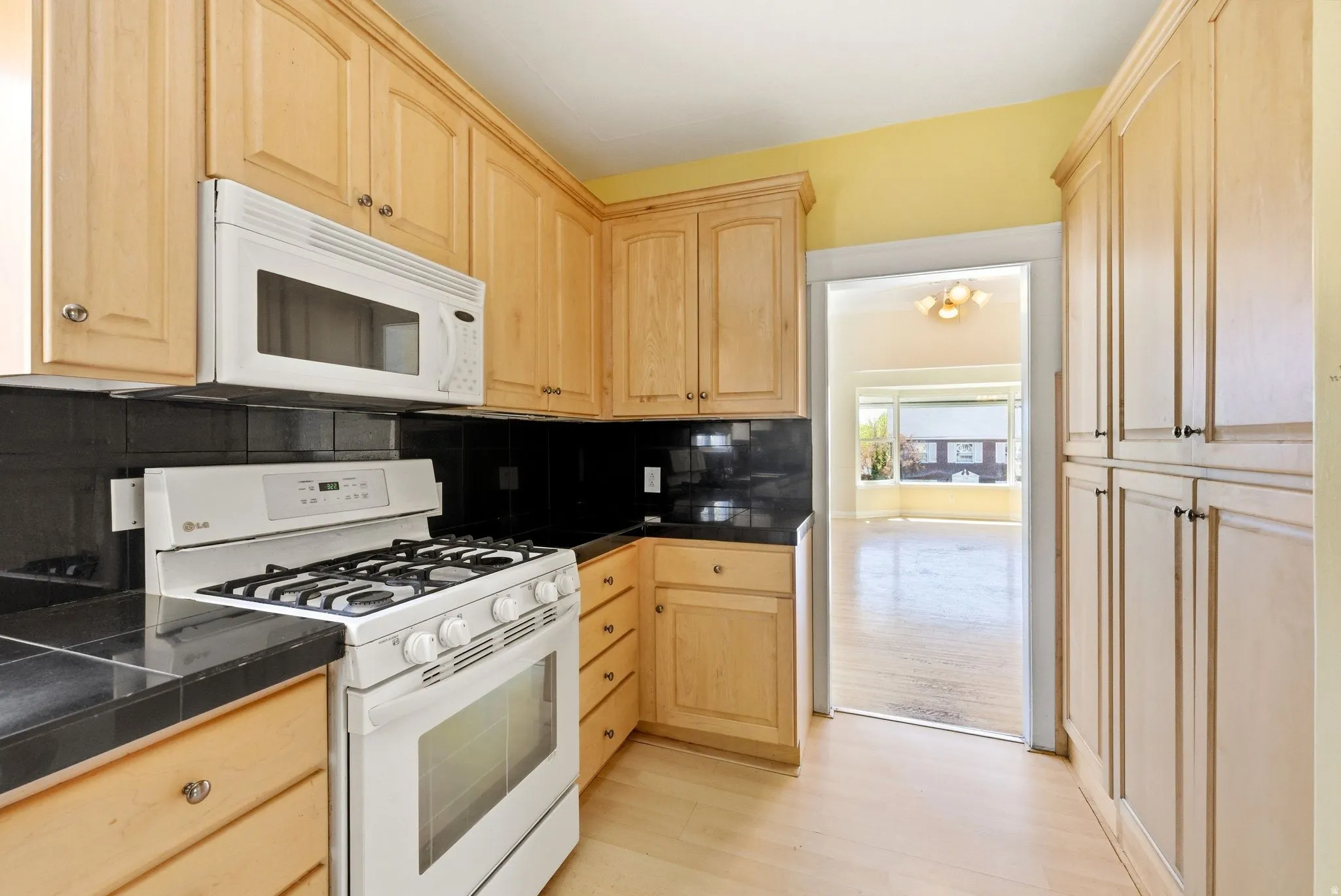 Kitchen with white appliances, light wood finish cabinetry, light wood-style flooring, decorative backsplash, and tile countertops
