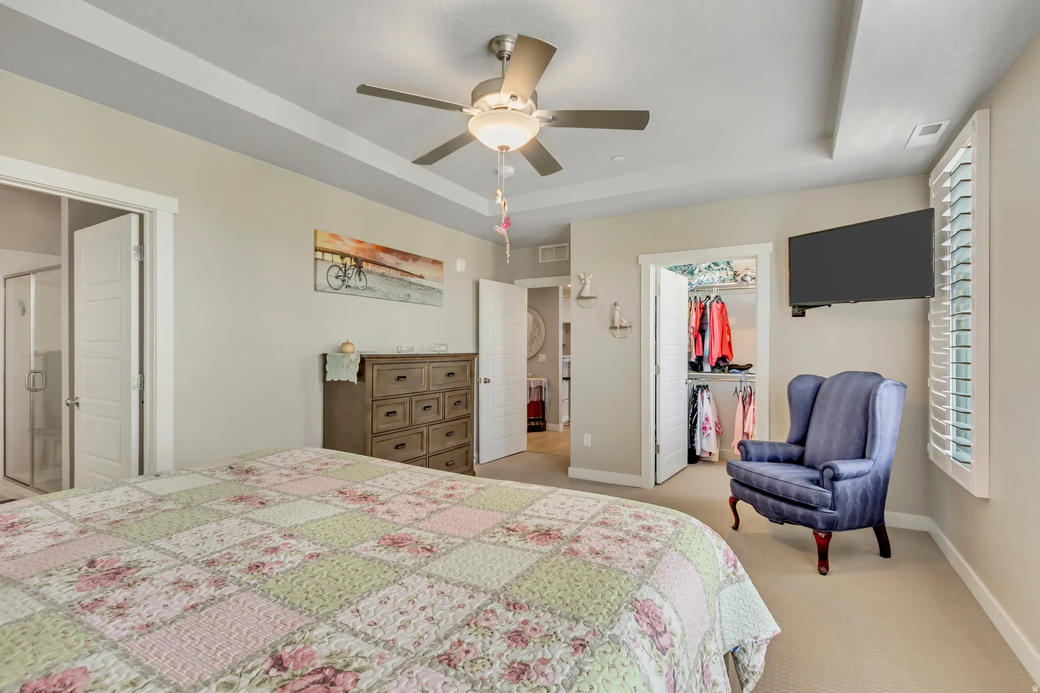 Bedroom featuring a tray ceiling, a walk in closet, light carpet, a ceiling fan, and ensuite bathroom