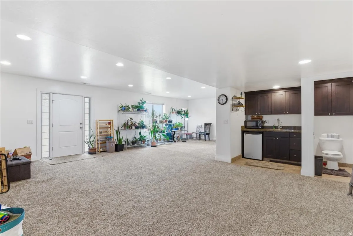 Indoor wet bar with light colored carpet, dark wood finish cabinets, refrigerator, recessed lighting, and dark countertops
