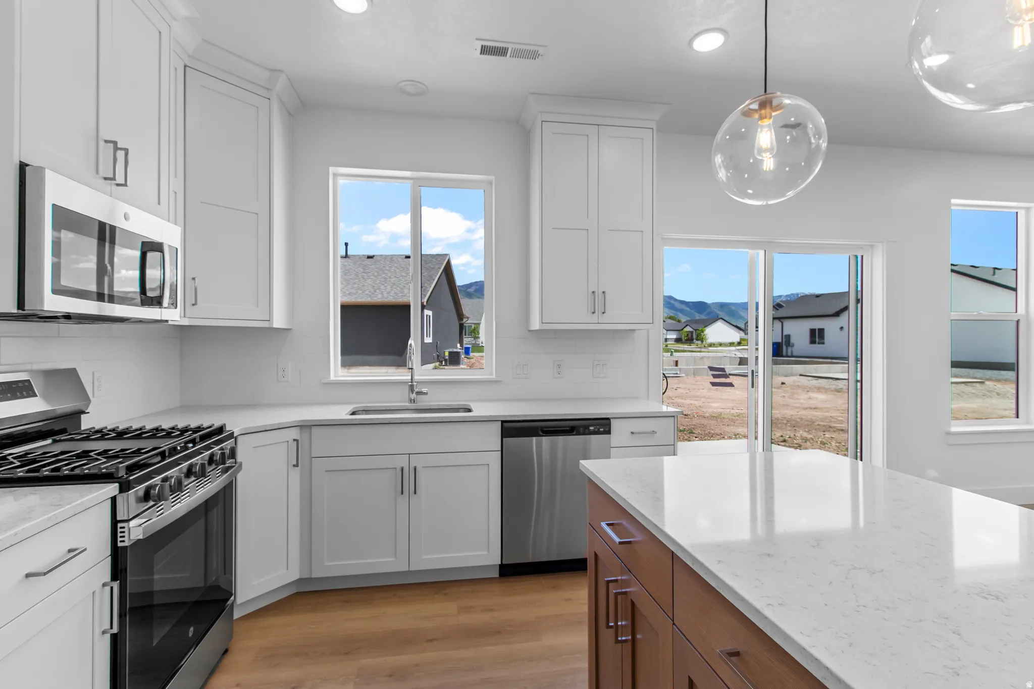 Kitchen with stainless steel appliances, two tone cabinetry, light wood-type flooring, pendant lighting, and light stone countertops