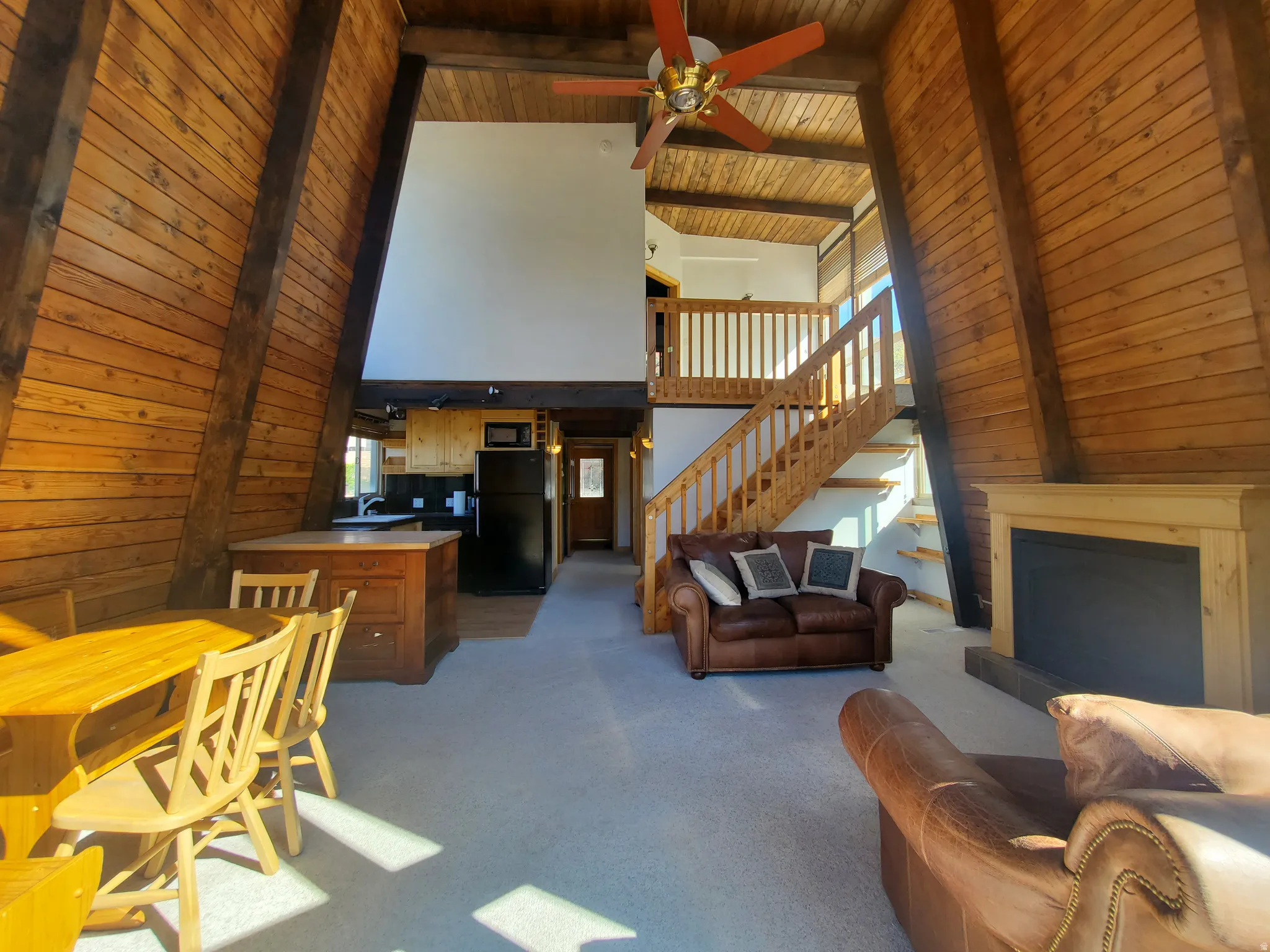Living room with carpet floors, a fireplace with raised hearth, a ceiling fan, a high wooden beamed ceiling, and wood walls