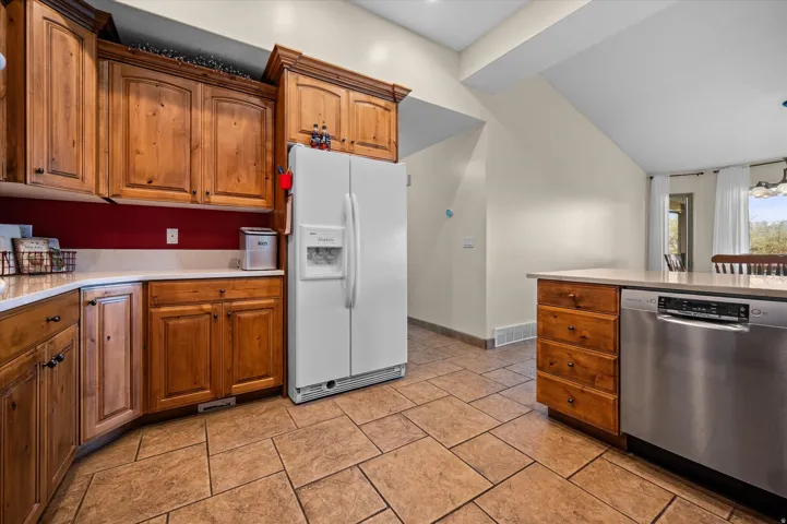 Kitchen with stainless steel dishwasher, white refrigerator with ice dispenser, wood finish cabinets, lofted ceiling, and light stone countertops