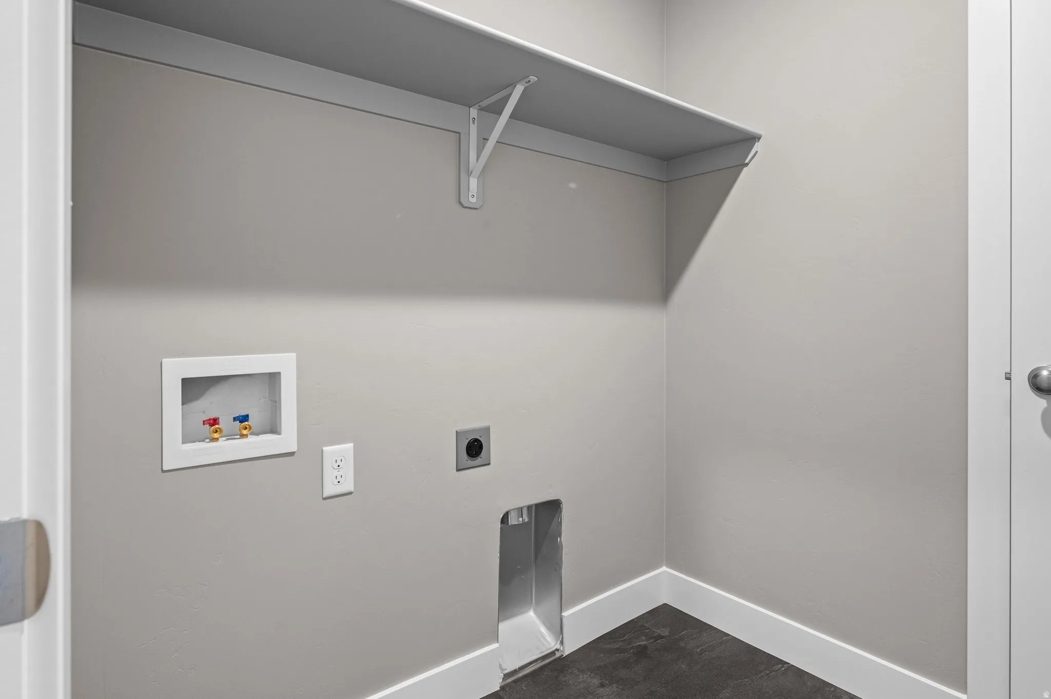 Dedicated utility space featuring a built-in shelf, wall-mounted water hookups, electrical outlets, and dark tile flooring