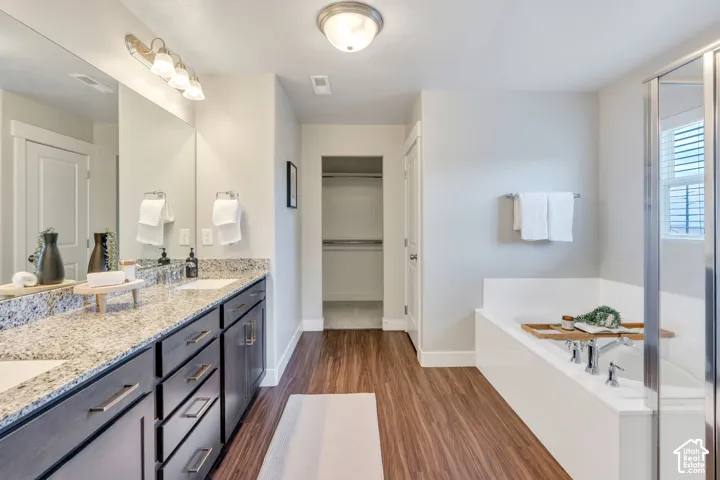 Dual vanity with stone countertop, wood-finish flooring, built-in tub, separate shower enclosure, and recessed closet