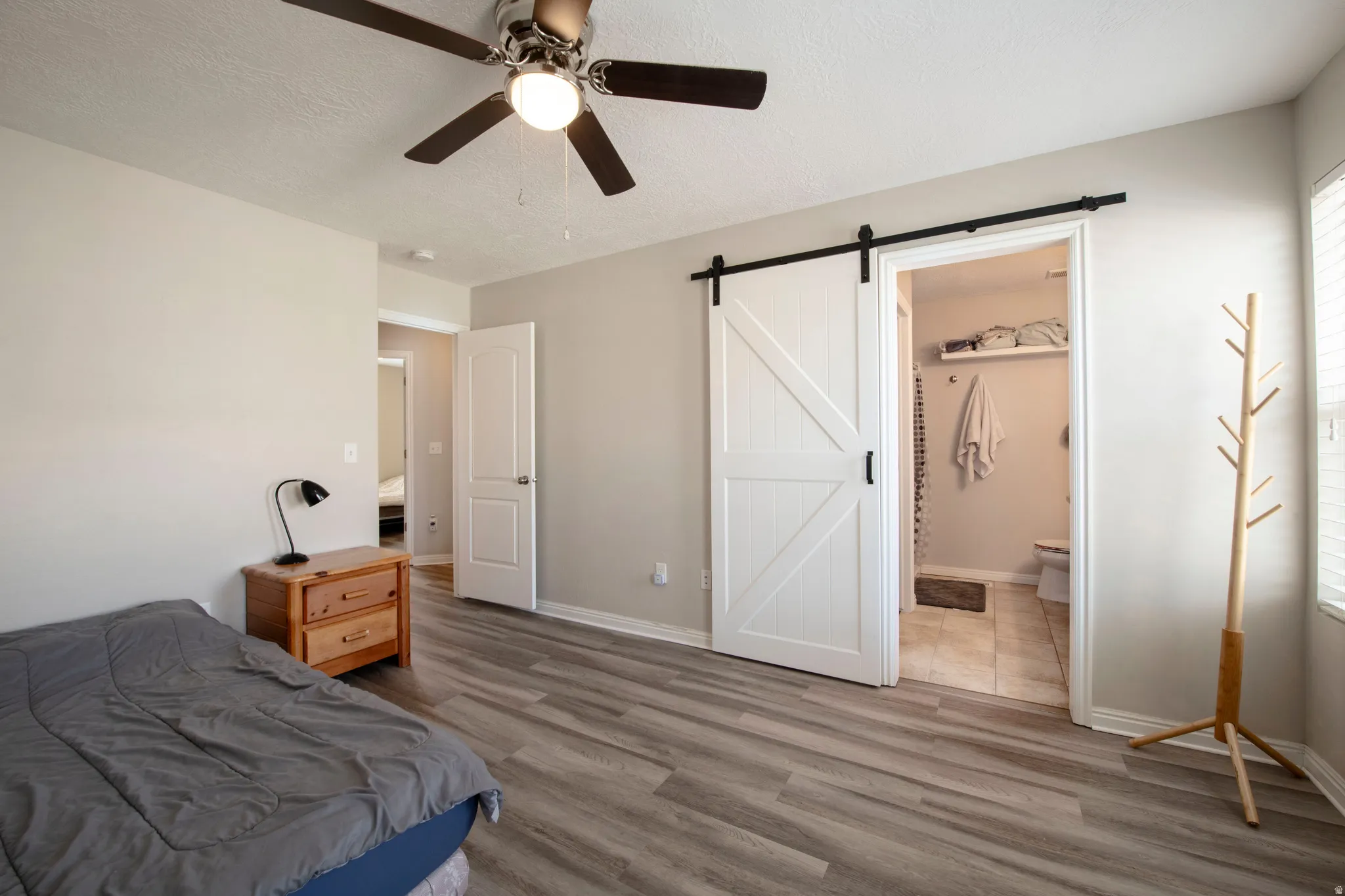 Bedroom featuring a barn door, light wood-style flooring, a textured ceiling, a ceiling fan, and ensuite bathroom