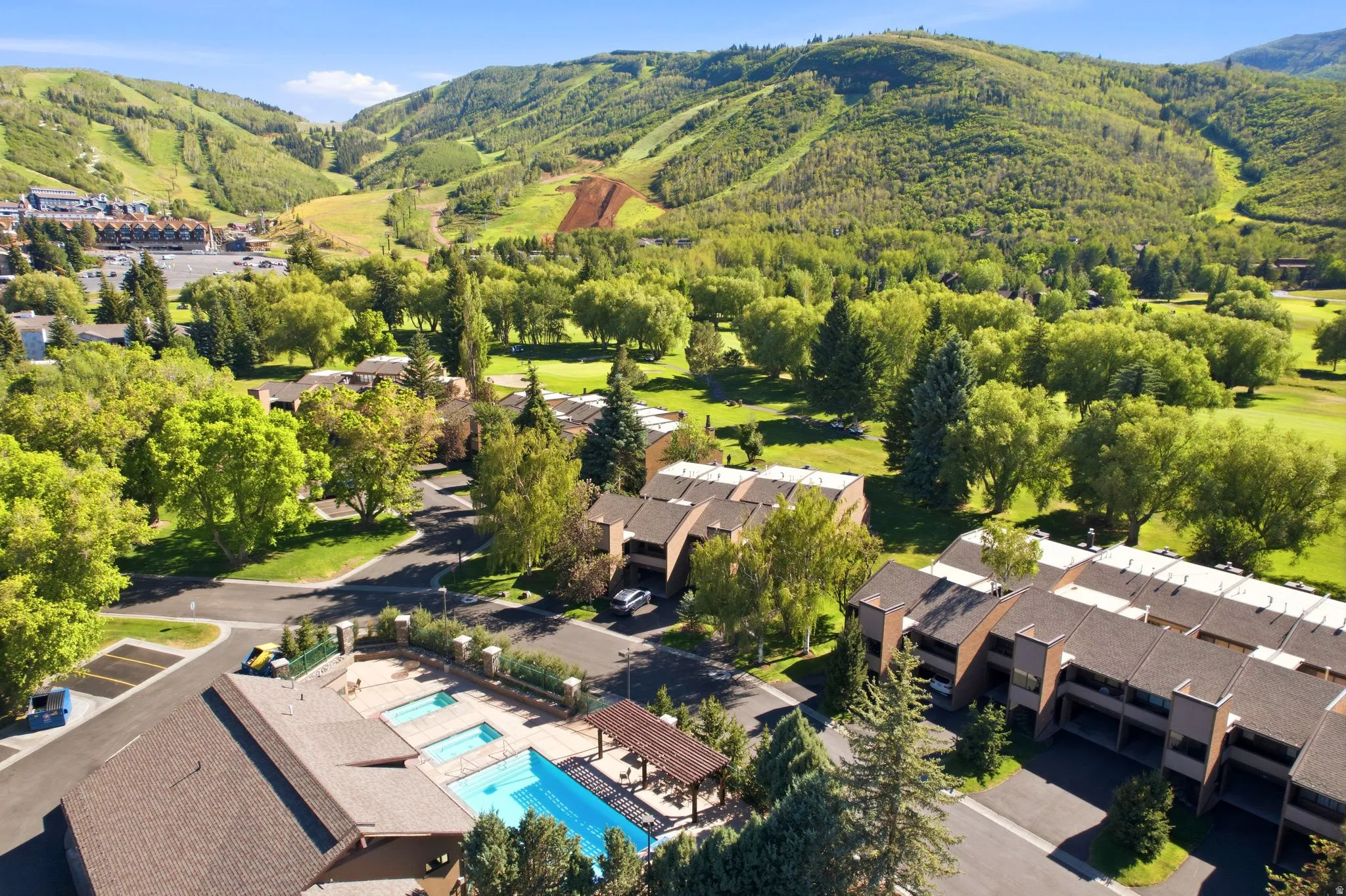 Resort-style outdoor pool area featuring multiple pools, a shaded pergola, and surrounding lush landscaping