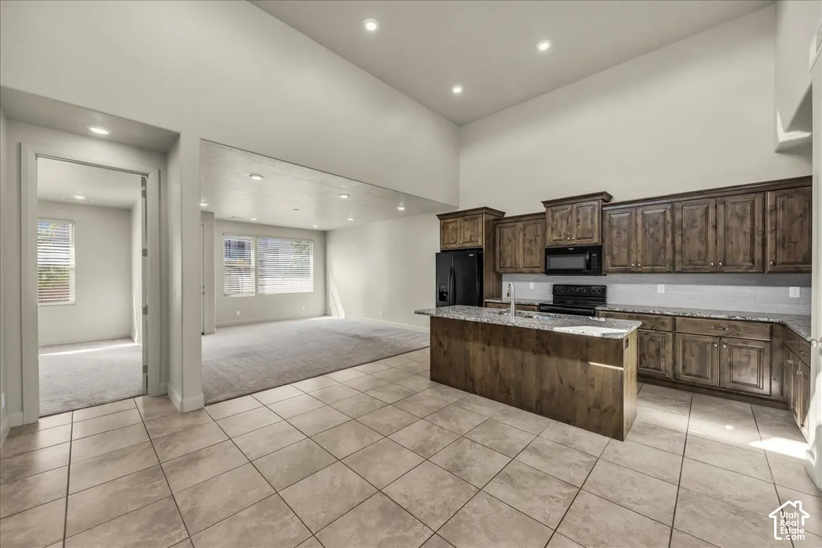 Kitchen with light tile patterned floors, an island with sink, light stone countertops, light colored carpet, and black appliances