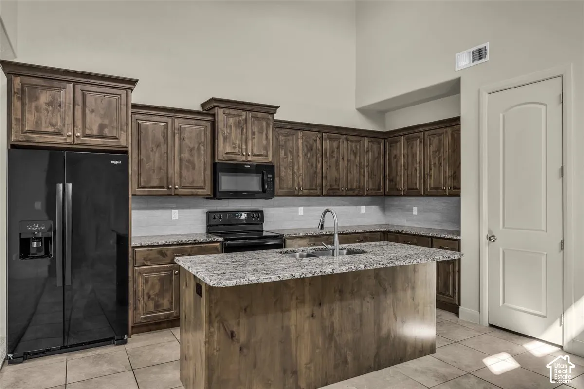 Kitchen with black appliances, dark wood finish cabinetry, light tile patterned flooring, a center island with sink, and light stone countertops