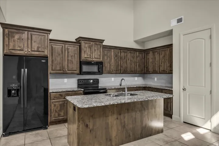 Kitchen with black appliances, dark wood finish cabinetry, light tile patterned flooring, a center island with sink, and light stone countertops