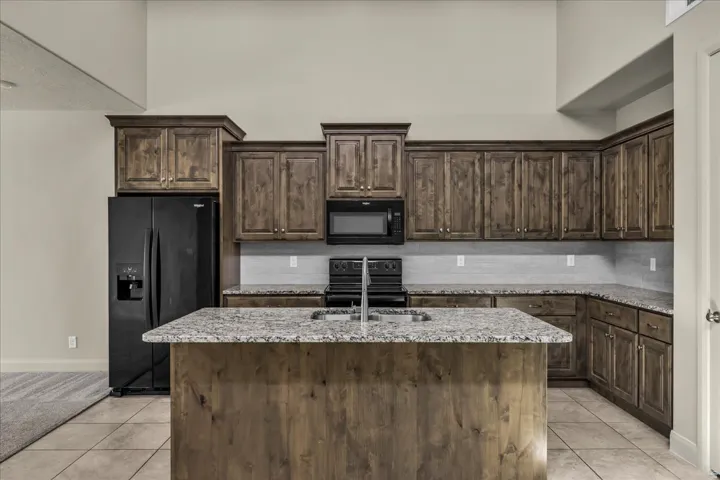 Kitchen featuring dark wood finish cabinets, black appliances, light stone counters, an island with sink, and light tile patterned floors