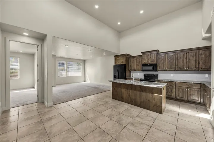 Kitchen with light tile patterned floors, an island with sink, light stone countertops, light colored carpet, and black appliances