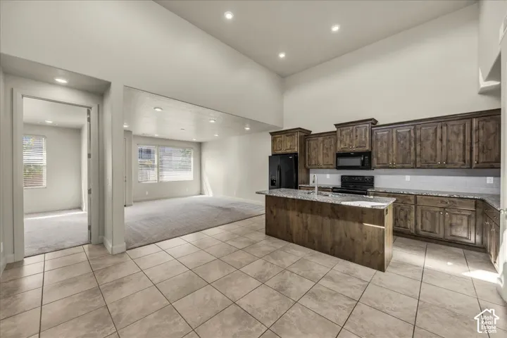 Kitchen with light tile patterned floors, an island with sink, light stone countertops, light colored carpet, and black appliances