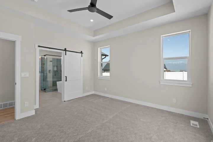 Unfurnished bedroom featuring a barn door, light carpet, a raised ceiling, ceiling fan, and ensuite bath