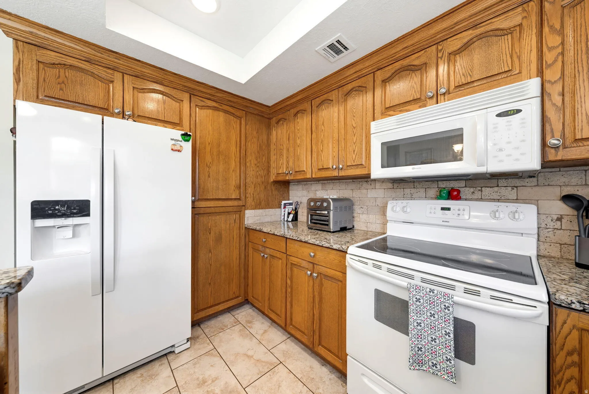 Kitchen featuring white appliances, wood finish cabinets, light stone countertops, tasteful backsplash, and a raised ceiling
