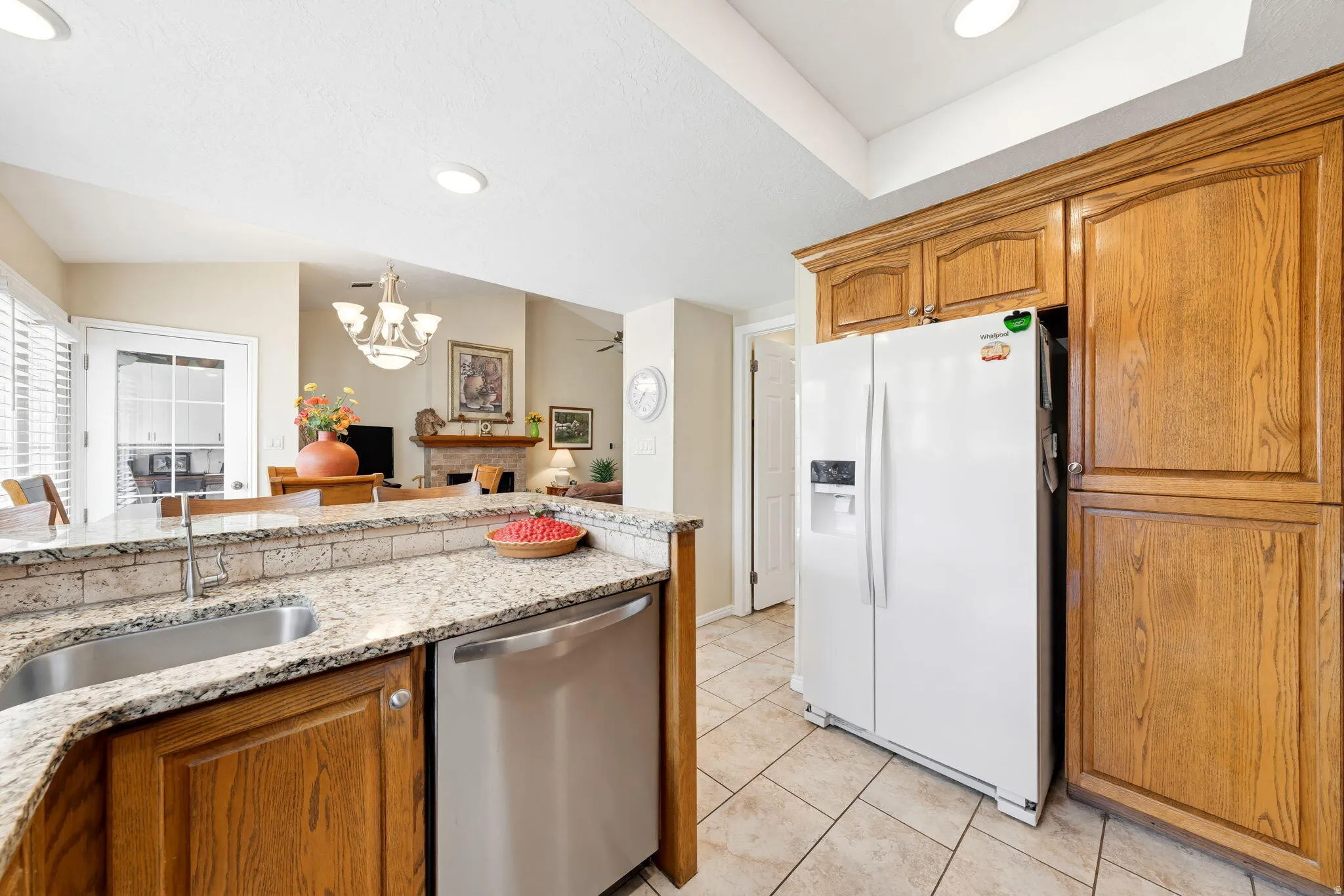 Kitchen with white refrigerator with ice dispenser, dishwasher, light stone countertops, wood finish cabinets, and hanging lights