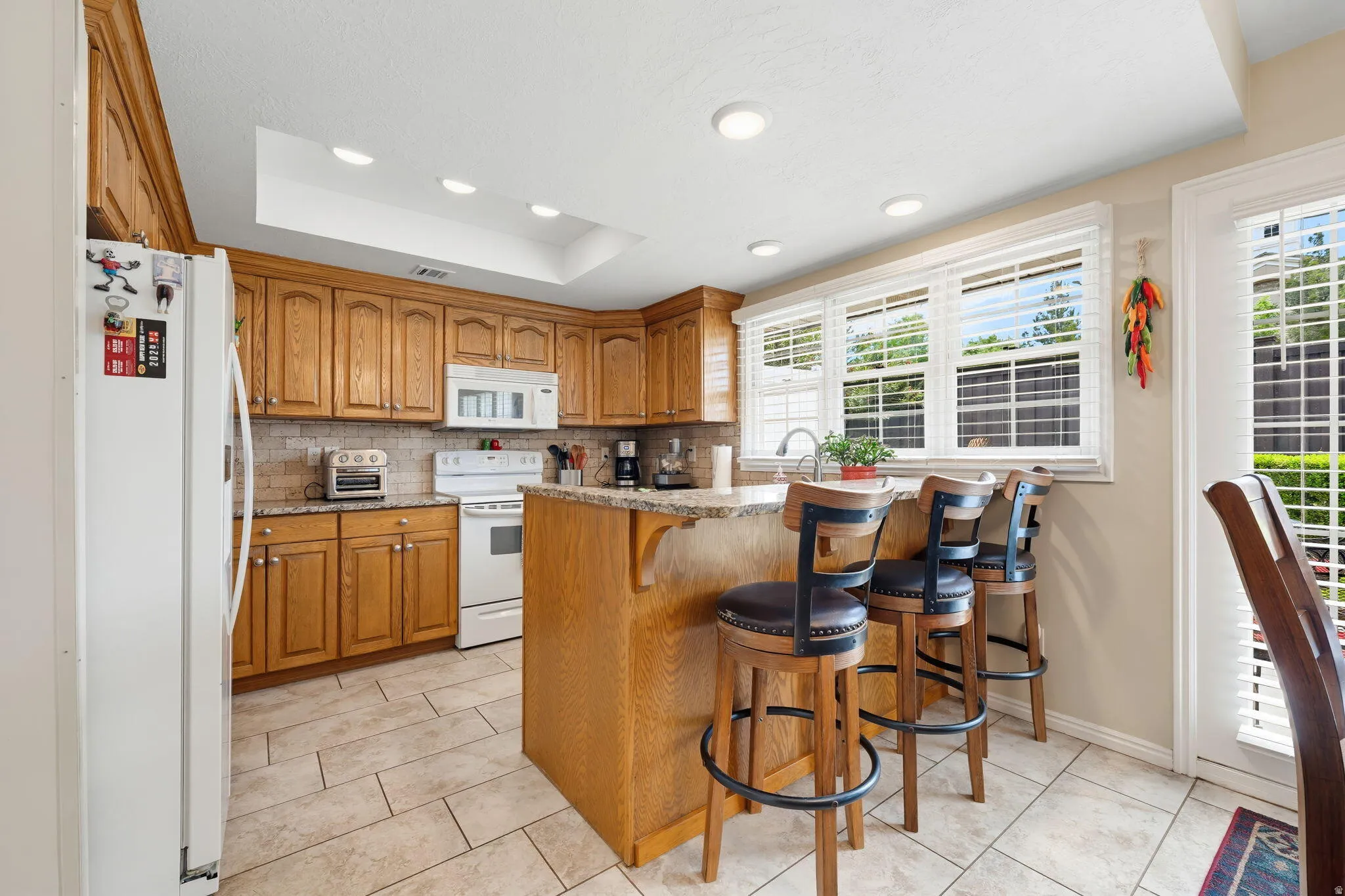 Kitchen featuring wood finish cabinetry, a kitchen bar, white appliances, light tile patterned flooring, and tasteful backsplash