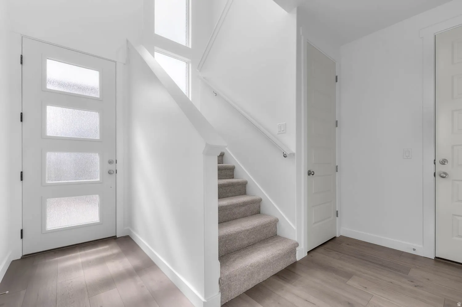 Entryway featuring a contemporary front door with frosted glass panels, wood-finish flooring, white walls, and a carpeted staircase with a modern handrail