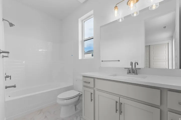 Bathroom featuring a white subway tile tub surround, white ceramic toilet, light gray vanity with an integrated sink, polished chrome fixtures, and a large mirror with a brass-finish vanity light