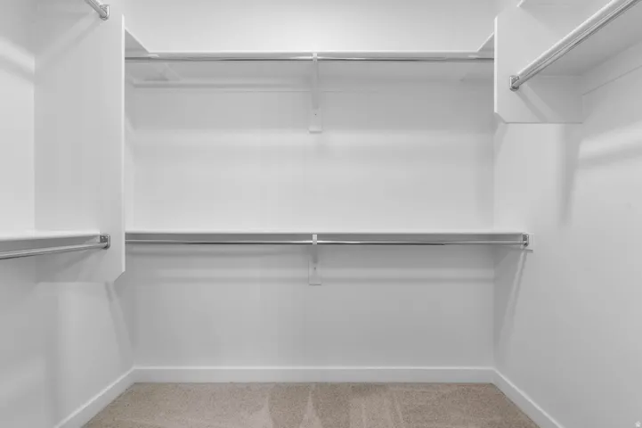Walk-in closet featuring white walls, white baseboards, multiple chrome hanging rods, and light-toned carpet flooring