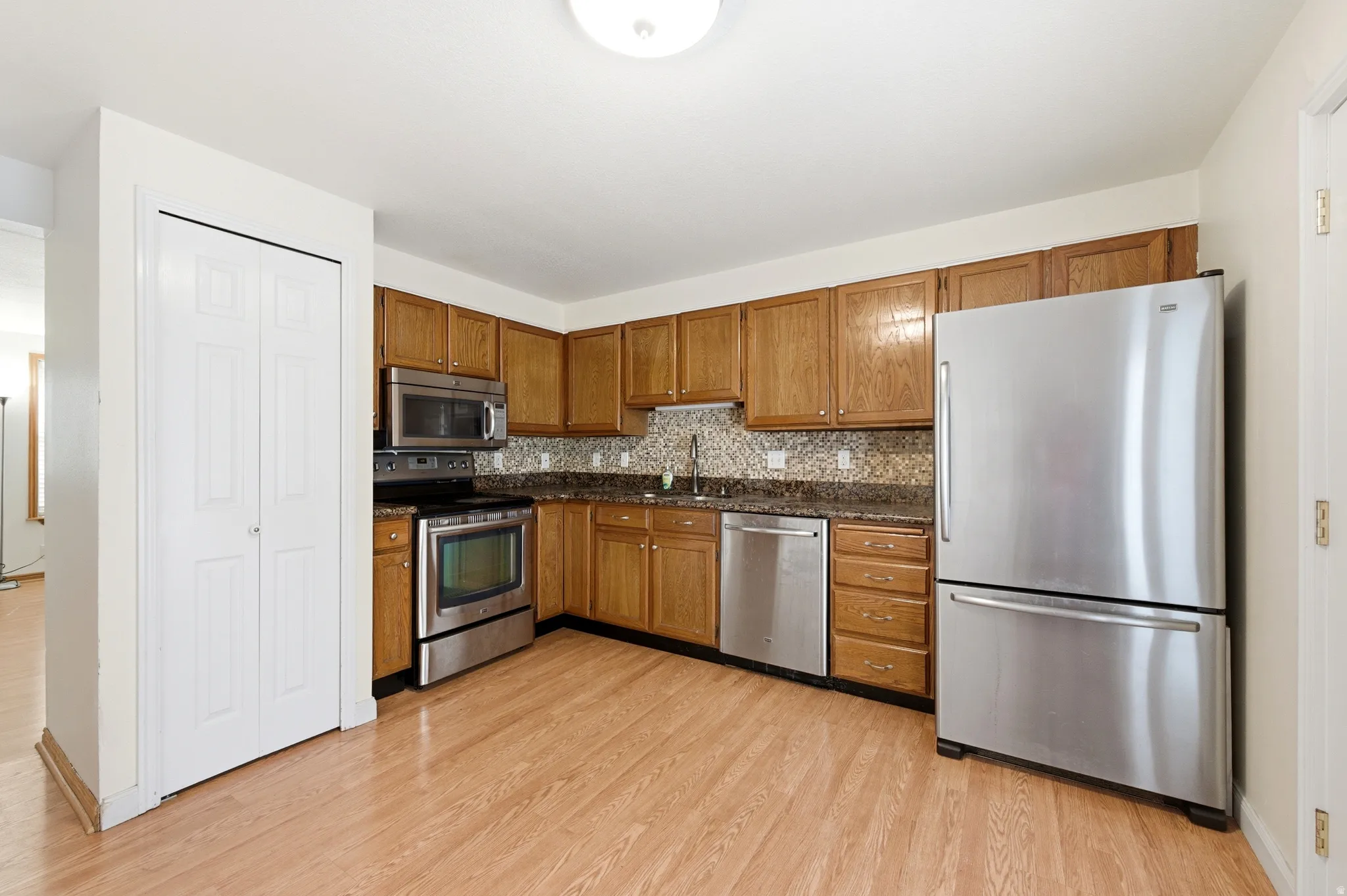 Kitchen with stainless steel appliances, wood finish cabinets, tasteful backsplash, light wood-type flooring, and dark stone countertops