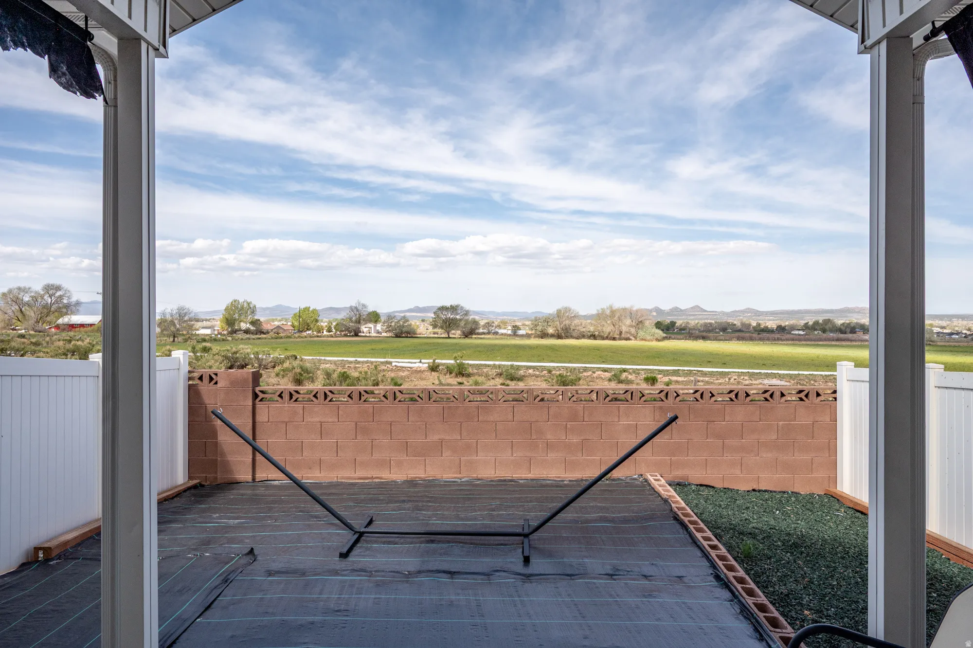 Covered patio with a dark ground covering, a partial cinder block wall with decorative top, a white vinyl fence, and a distant field