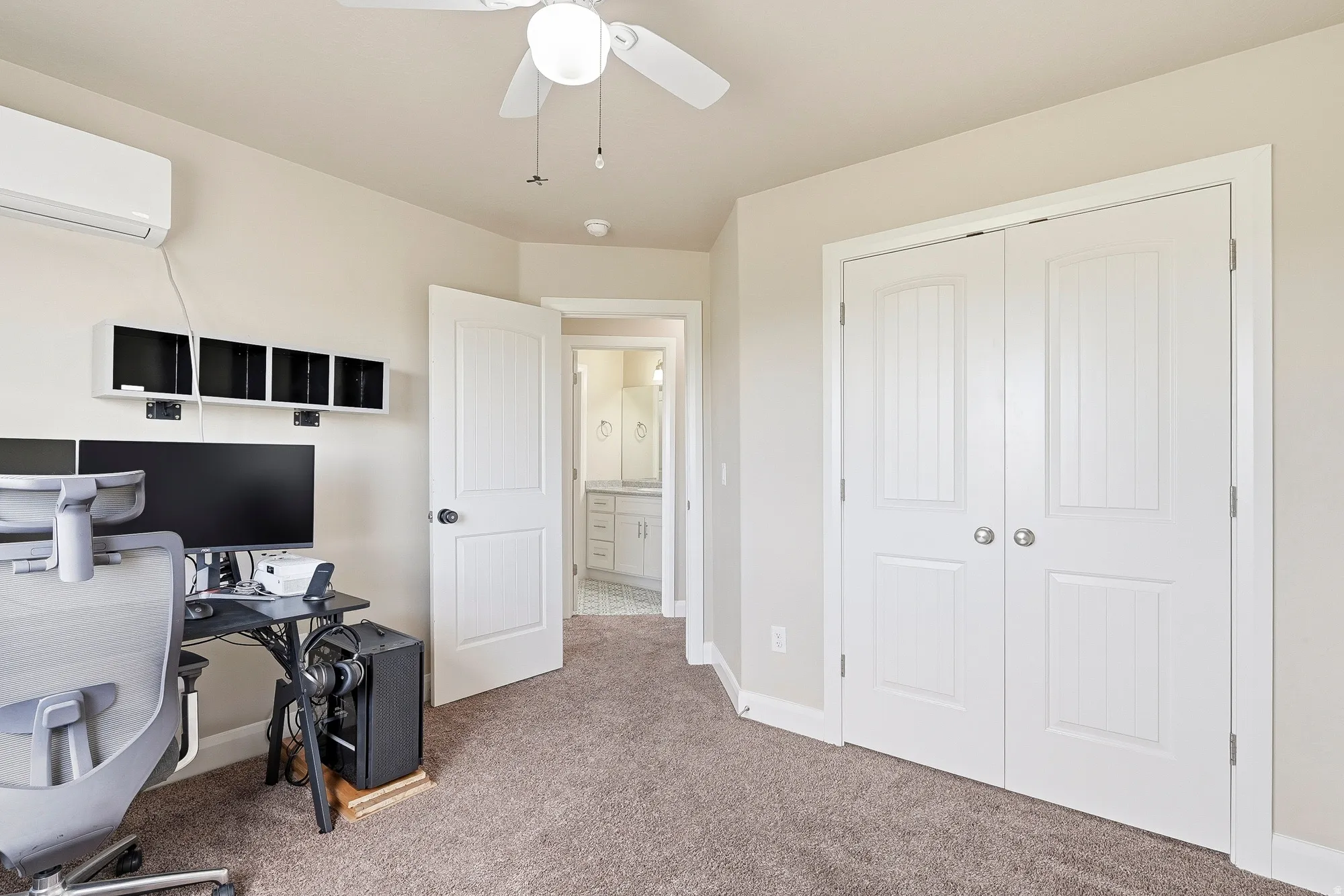 Plush carpeted room featuring a ceiling fan, white bi-fold closet doors, and an open doorway revealing a bathroom with a white vanity and tiled flooring