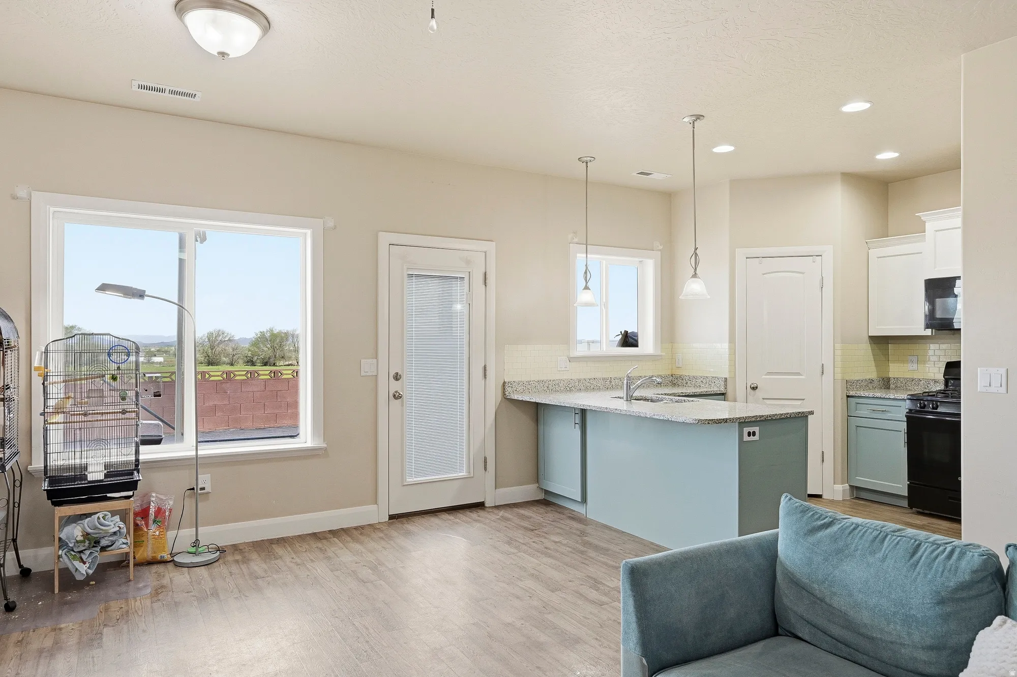 Open-concept living space featuring wood-finish flooring, a kitchen island with pendant lighting, white cabinetry, integrated black appliances, and a glass-paneled exterior door