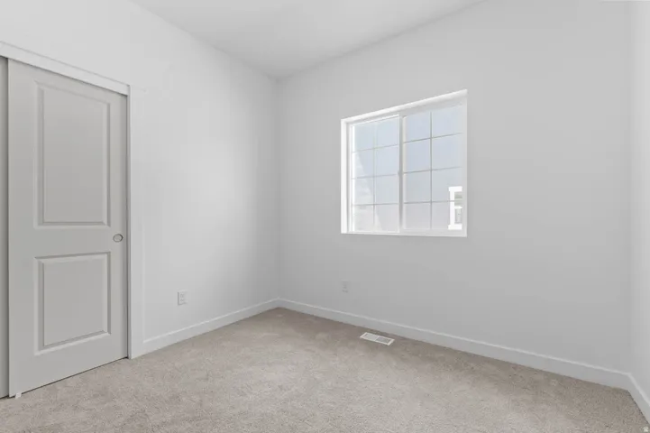 Bright interior room featuring a window with grid panes, neutral-toned carpeting, white walls, and a sliding door with a panel design