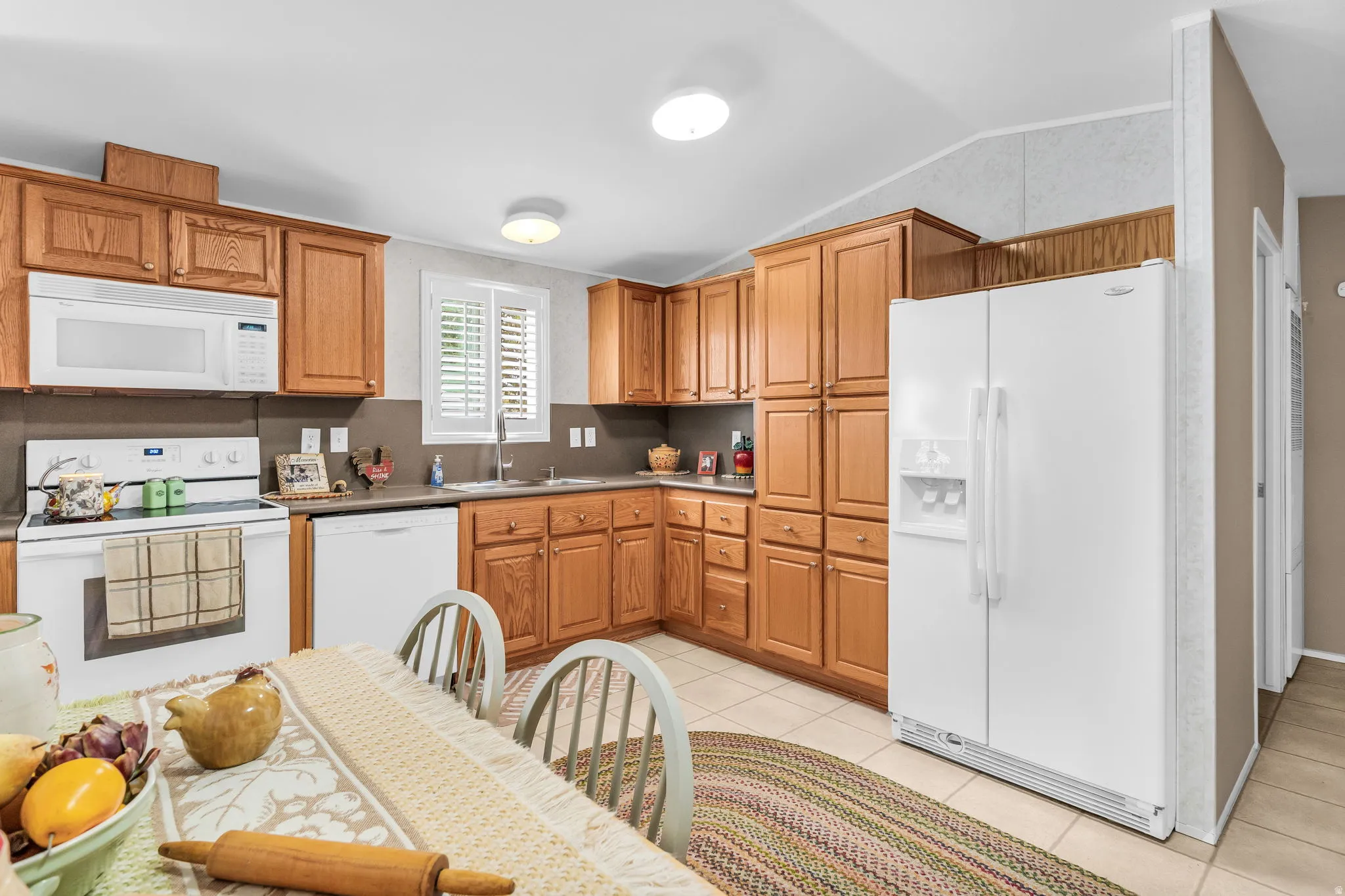 Kitchen with white appliances, light tile patterned flooring, light countertops, wood finish cabinetry, and vaulted ceiling