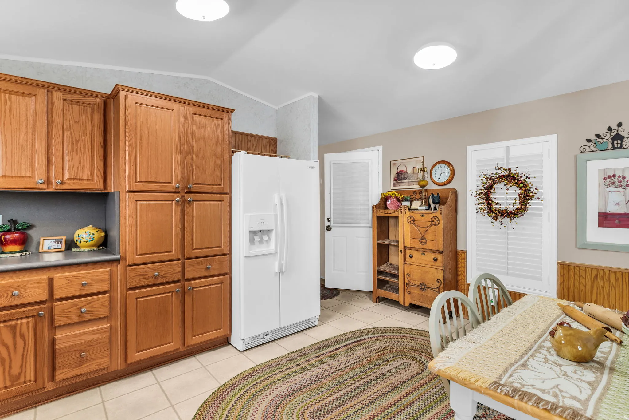 Kitchen featuring white refrigerator with ice dispenser, wood finish cabinets, light tile patterned floors, a wainscoted wall, and vaulted ceiling